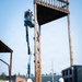 Military personnel in camouflage uniforms and white hard hats are conducting a construction or demolition operation on a large wooden tower structure. Two workers are elevated in a blue boom lift (aerial work platform) performing work at height, while three others observe from ground level. The setting appears to be a military installation based on the uniforms, vehicles, and surrounding buildings.