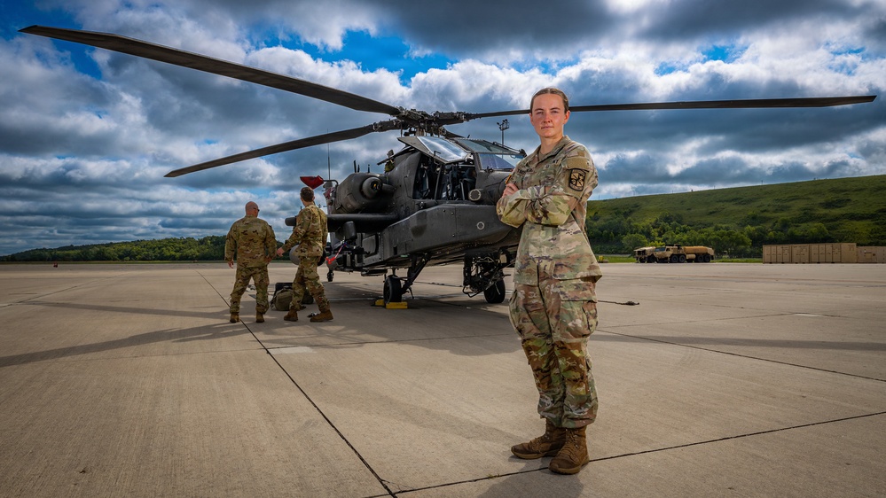 A U.S. Army soldier in Operational Camouflage Pattern (OCP) uniform stands with arms crossed in the foreground on a military airfield tarmac, posing in front of what appears to be an AH-64 Apache attack helicopter. Two additional soldiers are visible in the background near the aircraft. The setting appears to be a military installation with rolling green hills and a dramatic cloudy sky in the background.