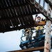 A construction worker is standing in a blue aerial lift (scissor lift or boom lift) platform, working on or inspecting an aged wooden overhead structure. The worker is equipped with full personal protective equipment including a hard hat, safety glasses, and a fall arrest harness. The photo is taken from a low angle looking upward, emphasizing the height and the weathered wooden roof structure above.
