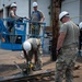 A group of five military personnel in camouflage uniforms and white hard hats are engaged in construction or maintenance work outdoors. Two individuals are positioned on a blue aerial lift platform, while three others work at ground level near wooden beams and utility poles. One person is using a power tool on lumber while others observe and assist.