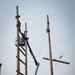 A worker in a blue aerial lift (boom lift) is dismantling or removing crossarms from tall wooden utility poles in what appears to be a military or industrial facility. A crossarm is visibly falling or being thrown from the right pole, captured mid-air during the removal process. The setting includes industrial buildings, a chain-link fence, and additional equipment in the background.