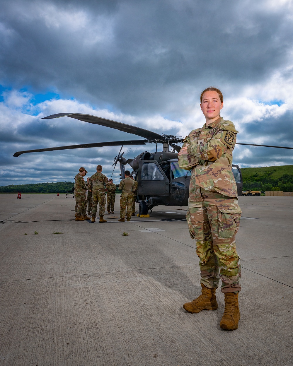 A female soldier in U.S. Army OCP (Operational Camouflage Pattern) uniform stands in the foreground on a flight line apron with arms crossed, posing for a portrait-style photograph. Behind her, a UH-60 Black Hawk helicopter is parked on the tarmac with a group of soldiers gathered around it, apparently conducting pre-flight or maintenance activities. The setting appears to be a military airfield with green hills and dramatic cloudy skies in the background.