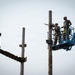 Two workers in military-style camouflage uniforms and white hard hats are elevated on a blue aerial lift (boom lift/cherry picker) working on a wooden utility pole. One worker appears to be operating a chainsaw or cutting tool against the pole. The scene appears to involve demolition or maintenance of an older wooden structure with multiple poles and cross-members visible.