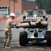A military service member wearing camouflage uniform and white hard hat is handling a large log loaded on a forklift or telehandler in a paved parking area. The scene appears to be a construction or engineering training exercise near a brick building with active construction visible in the background. Multiple pieces of heavy equipment and vehicles are present in the area.