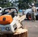 A STIHL chainsaw rests on a freshly cut tree stump in the foreground, with military or utility personnel working in the background. A Bobcat skid-steer loader (model 863) is visible behind one worker wearing a white hard hat, suggesting tree removal or debris clearing operations. The scene appears to take place in a paved lot near a building, likely during a post-storm or routine grounds maintenance operation.