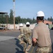 Military personnel in camouflage uniforms and white hard hats are engaged in what appears to be a construction or engineering training exercise at a military installation. Several soldiers are pulling a yellow rope or cable toward a structure, while a supervisor or safety officer in a plain tan shirt and white hard hat observes from behind. The setting includes a paved area with construction infrastructure, vehicles, and tree-lined surroundings consistent with a U.S. military base.
