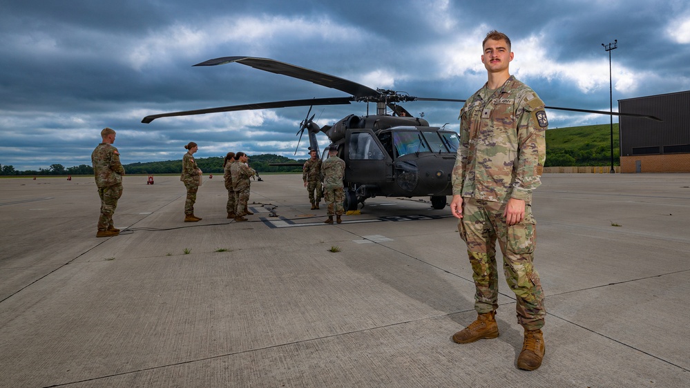 A U.S. Army soldier stands in the foreground on a military airfield tarmac, posing for what appears to be an official or promotional photograph. Behind him, several other soldiers in OCP (Operational Camouflage Pattern) uniforms are gathered around a UH-60 Black Hawk helicopter. The scene is set under a dramatic, overcast sky with green hills visible in the background.