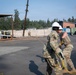 Two military personnel in camouflage uniforms are actively handling a yellow rope or cable on a paved area, with one wearing a white hard hat. The scene appears to be a training or work exercise at a military installation or industrial facility. A third uniformed individual is visible in the background near a barrier wall.