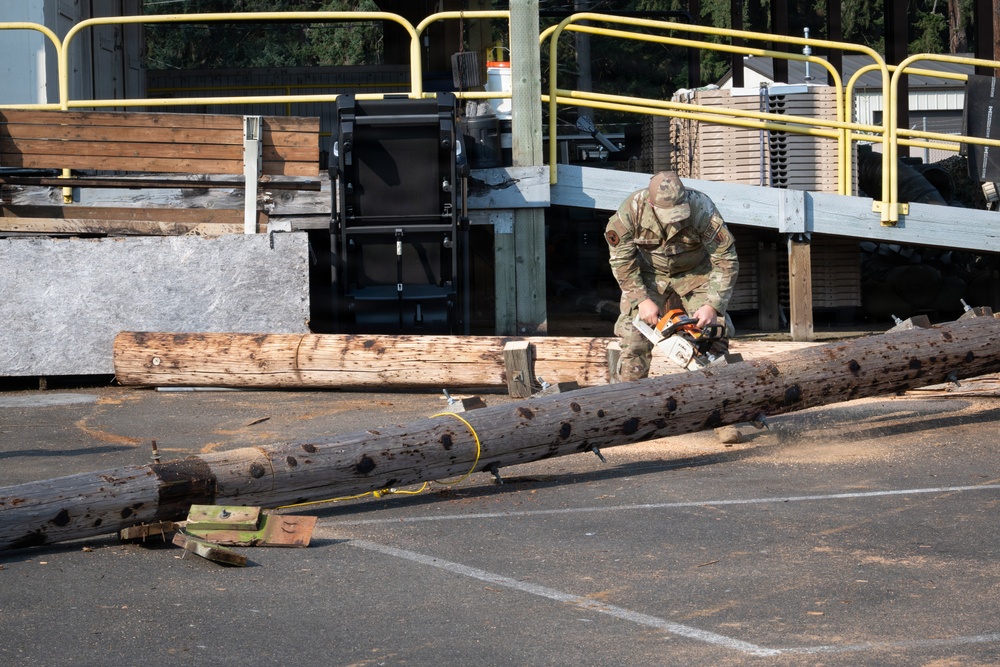 A person in military camouflage uniform is using an orange-and-white chainsaw to cut through a fallen utility pole or large log on a paved surface. The scene appears to be a debris clearance operation, likely following a storm or natural disaster. The setting includes a loading dock area with yellow safety railings and stacked materials in the background.