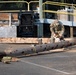 A person in military camouflage uniform is using an orange-and-white chainsaw to cut through a fallen utility pole or large log on a paved surface. The scene appears to be a debris clearance operation, likely following a storm or natural disaster. The setting includes a loading dock area with yellow safety railings and stacked materials in the background.