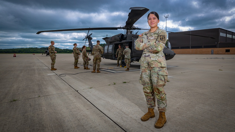 A female U.S. Army soldier stands in the foreground on a tarmac, posing with arms crossed and smiling at the camera. Behind her, a group of soldiers in OCP (Operational Camouflage Pattern) uniforms are gathered around what appears to be a UH-60 Black Hawk helicopter near a military hangar. The scene is set under overcast, dramatic skies at what appears to be a military airfield.