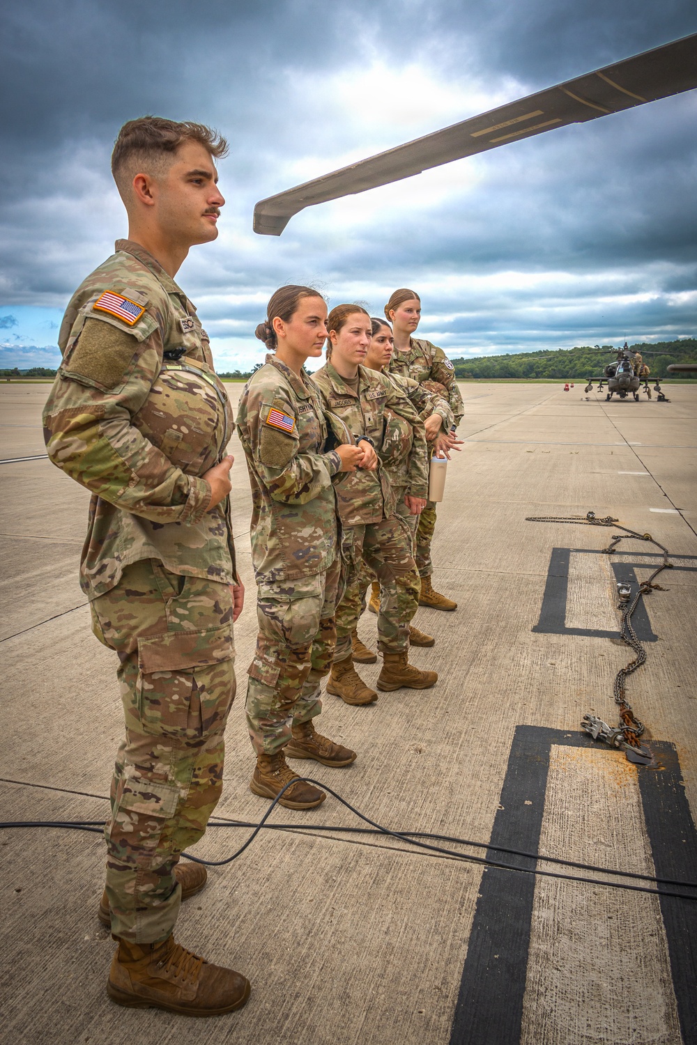A group of U.S. Army soldiers in OCP (Operational Camouflage Pattern) uniforms stand on a flight line or tarmac, appearing to receive a briefing or instruction. An Apache attack helicopter (AH-64) is visible in the background on the tarmac. The setting appears to be a military airfield during overcast conditions.