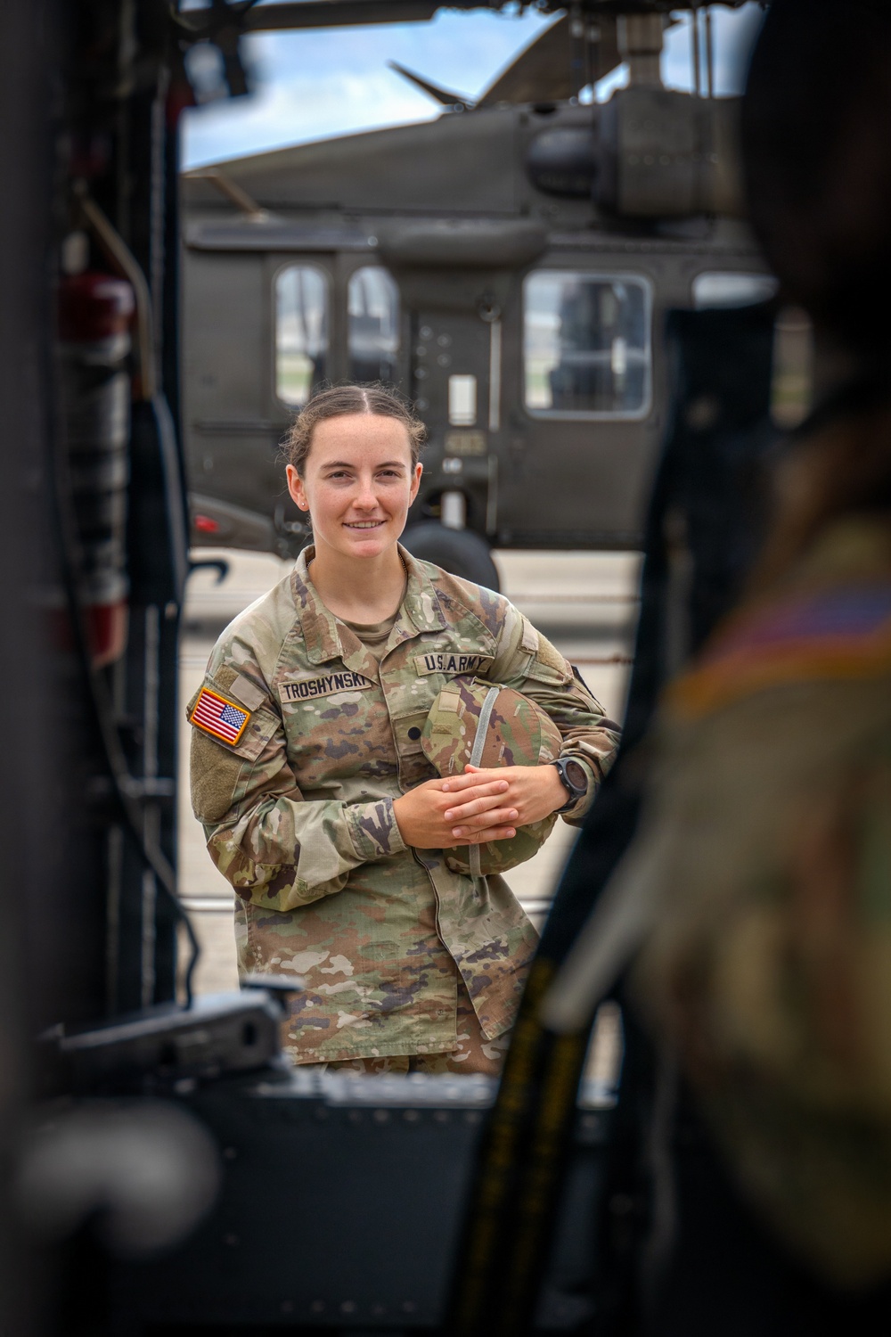 A U.S. Army soldier named Troshynski, wearing Operational Camouflage Pattern (OCP) uniform, is photographed smiling on what appears to be a military airfield or flight line. The image is framed through the doorway or opening of a military helicopter, with another helicopter visible in the background. The composition suggests a candid or portrait-style military documentation photograph.