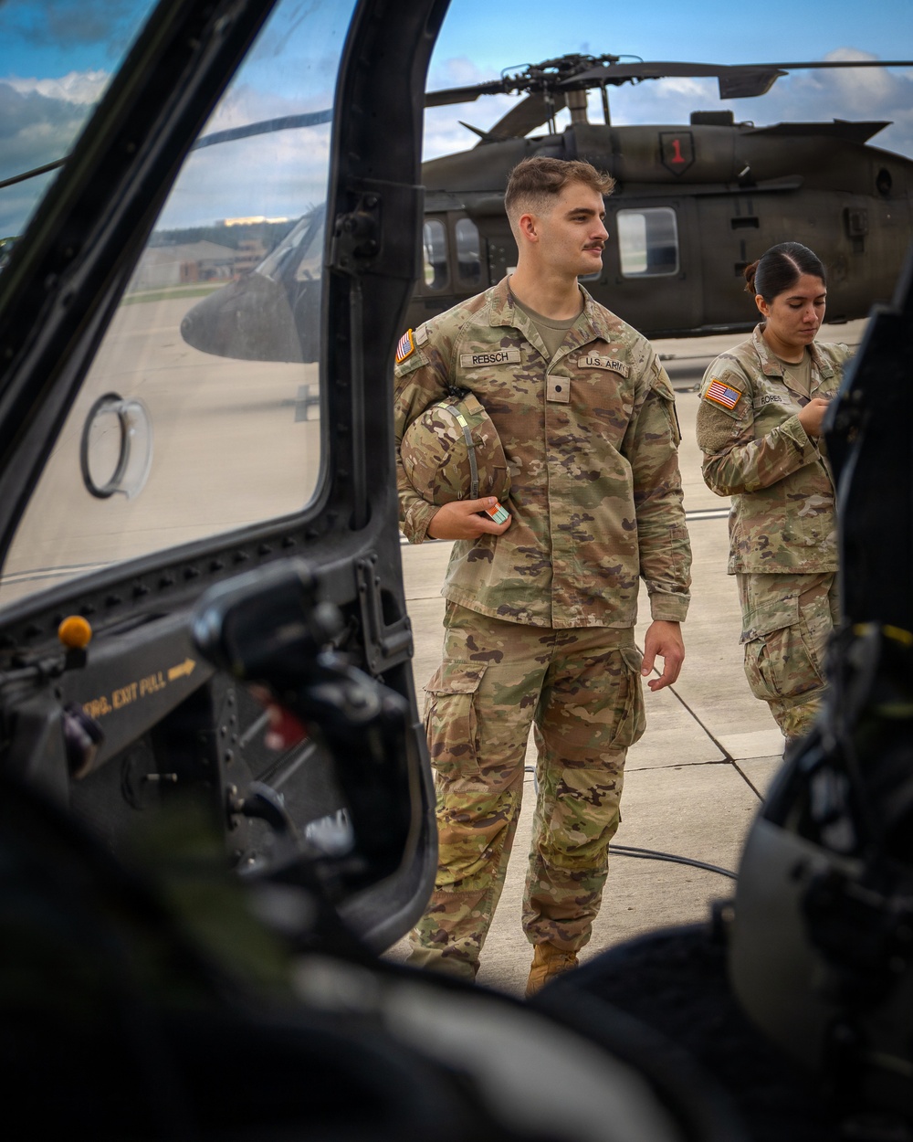 Two U.S. Army soldiers in OCP (Operational Camouflage Pattern) uniforms stand on a military flight line near helicopters. The foreground soldier, identified by nametape as 'REBSCH,' holds a flight helmet and appears to be conducting pre-flight or post-flight activities. A UH-60 Black Hawk helicopter marked with the number '1' is visible in the background.