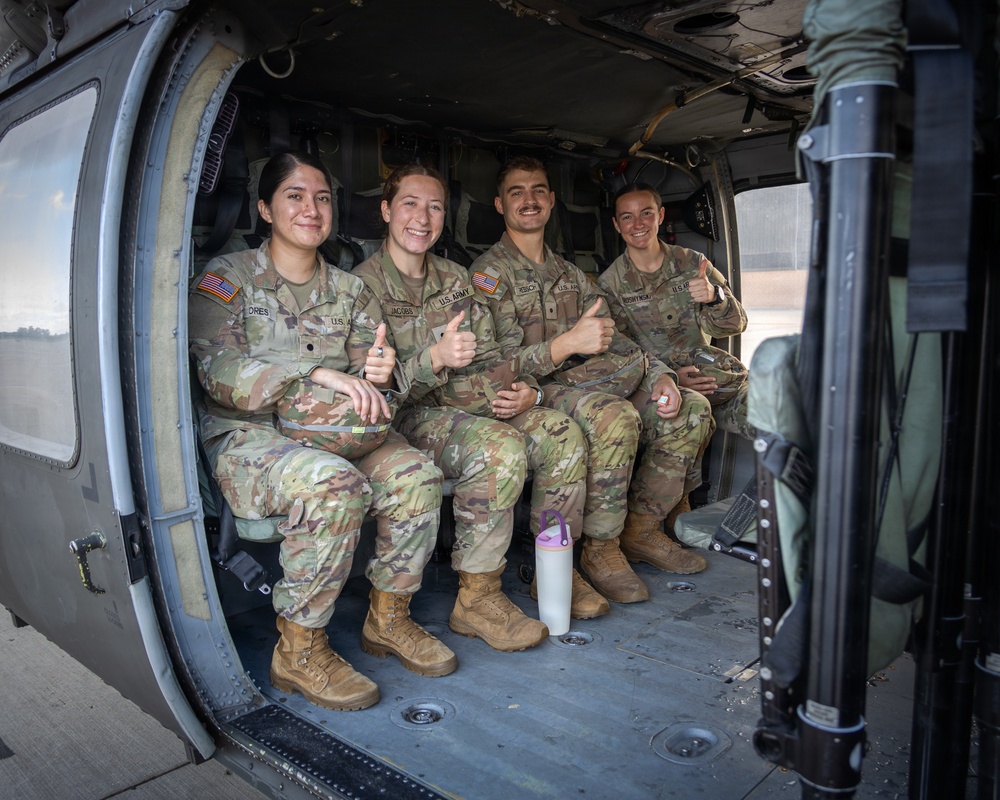 Four U.S. Army soldiers in OCP (Operational Camouflage Pattern) uniforms are seated inside what appears to be a UH-60 Black Hawk helicopter, giving thumbs-up gestures and smiling at the camera. Three appear to be female and one male, all wearing American flag patches and 'U.S. ARMY' tape on their uniforms. The image captures a candid, morale-positive moment likely during a training exercise or mission.