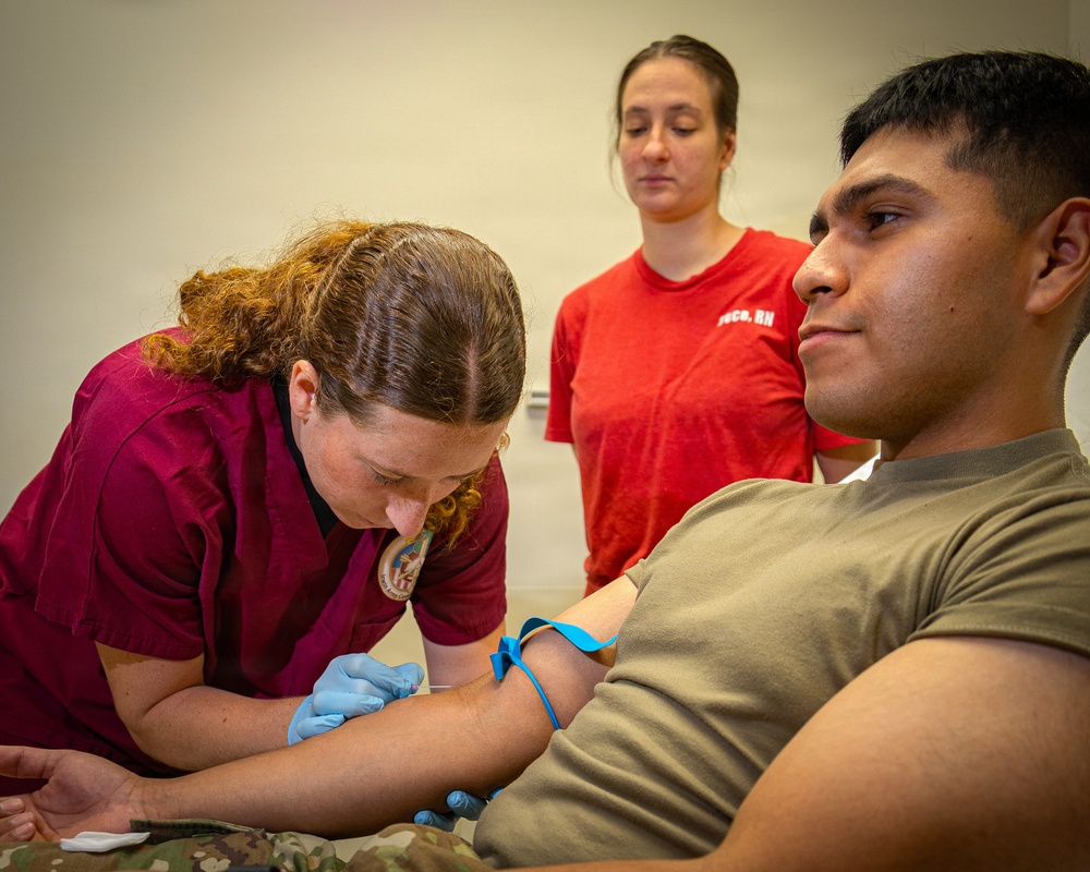 A medical professional in dark red scrubs wearing blue latex gloves is performing a venipuncture or blood draw on a young male patient wearing an olive/tan military-style t-shirt and camouflage pants. A blue tourniquet band is visible on the patient's arm. A second person in a red t-shirt with 'RN' text visible is observing the procedure in the background.