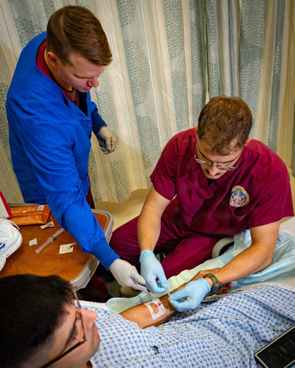 Two medical professionals wearing gloves are performing what appears to be an intravenous (IV) line insertion or wound care procedure on a patient lying in a hospital bed. One person in maroon scrubs with a medical patch is actively working on the patient's arm, while a second person in a blue lab coat assists. A side table with medical supplies including syringes, packaging, and a basin is visible nearby.