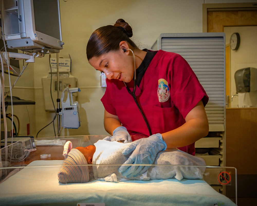 A healthcare worker in red scrubs with a stethoscope examines a newborn infant in a hospital bassinet. The setting appears to be a maternity ward or neonatal care unit, with medical equipment visible in the background. The caregiver is wearing blue latex gloves and appears attentive and engaged with the infant.