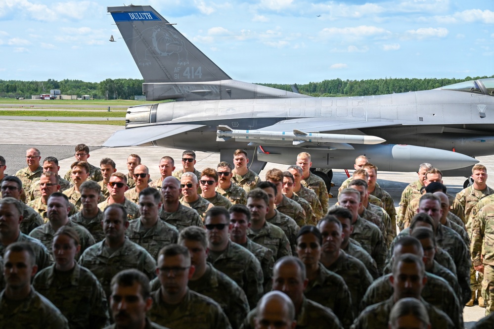A large formation of military personnel in camouflage uniforms are assembled on an airfield tarmac in front of a U.S. Air Force F-16 Fighting Falcon jet. The aircraft's tail fin displays the word 'DULUTH' in blue lettering along with the serial number 'AF 414', indicating it belongs to the Duluth-based 148th Fighter Wing, Minnesota Air National Guard. The scene appears to be a formal military ceremony or group photo event.