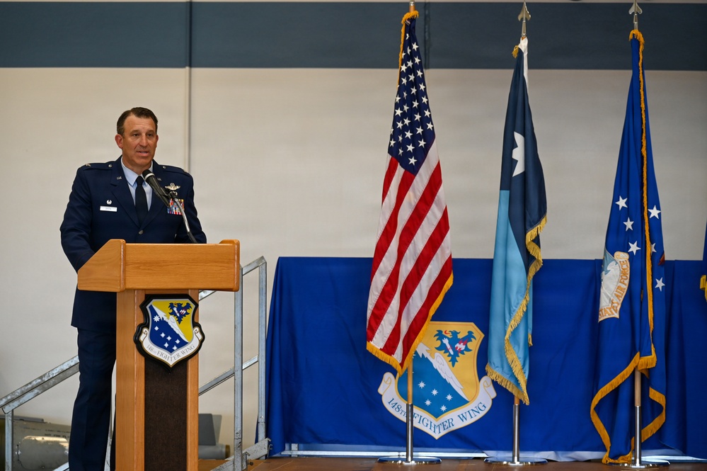 A U.S. Air Force officer in dress blues is speaking at a podium during an official ceremony for the 148th Fighter Wing. The setting appears to be a hangar or large indoor military facility, with the American flag and military unit flags displayed prominently behind the speaker. The backdrop features a blue drape with the 148th Fighter Wing emblem.