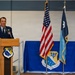 A U.S. Air Force officer in dress blues is speaking at a podium during an official ceremony for the 148th Fighter Wing. The setting appears to be a hangar or large indoor military facility, with the American flag and military unit flags displayed prominently behind the speaker. The backdrop features a blue drape with the 148th Fighter Wing emblem.