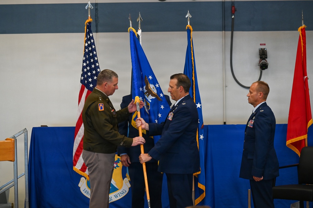 A military change of command ceremony is taking place, likely at the 148th Fighter Wing based on the visible emblem on the blue tablecloth. An Army officer in olive green uniform is exchanging a guidon/flag with an Air Force officer in dress blues, representing the formal transfer of command. A third Air Force officer stands at attention to the right as a witness to the ceremony.