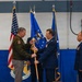 A military change of command ceremony is taking place, likely at the 148th Fighter Wing based on the visible emblem on the blue tablecloth. An Army officer in olive green uniform is exchanging a guidon/flag with an Air Force officer in dress blues, representing the formal transfer of command. A third Air Force officer stands at attention to the right as a witness to the ceremony.