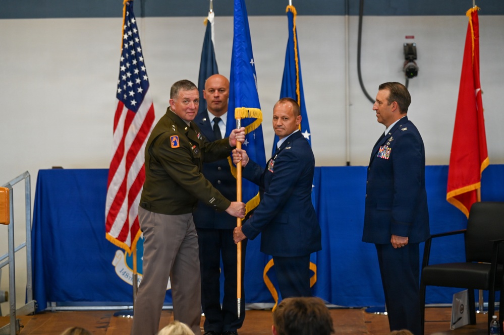 A military change of command ceremony is taking place in what appears to be a hangar or gymnasium decorated with blue draping. An Army officer in green uniform is passing or receiving a guidon (unit flag on a staff) to/from an Air Force officer in dress blues, signifying a transfer of command. Additional uniformed personnel stand at attention on the stage, with multiple flags including the US flag and Air Force flags visible in the background.