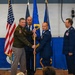 A military change of command ceremony is taking place in what appears to be a hangar or gymnasium decorated with blue draping. An Army officer in green uniform is passing or receiving a guidon (unit flag on a staff) to/from an Air Force officer in dress blues, signifying a transfer of command. Additional uniformed personnel stand at attention on the stage, with multiple flags including the US flag and Air Force flags visible in the background.