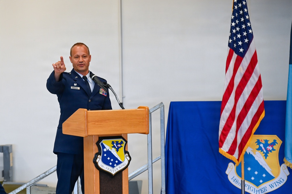 A U.S. Air Force officer in dress blues is speaking at a podium during an official ceremony for the 148th Fighter Wing. He is gesturing with his right hand raised, pointing upward, while addressing an audience. The setting appears to be inside a hangar or large facility decorated with unit insignia and an American flag.