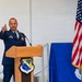 A U.S. Air Force officer in dress blues is speaking at a podium during an official ceremony for the 148th Fighter Wing. He is gesturing with his right hand raised, pointing upward, while addressing an audience. The setting appears to be inside a hangar or large facility decorated with unit insignia and an American flag.