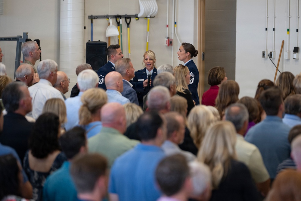 A military ceremony or event is taking place indoors, likely in a gymnasium or multipurpose facility. Three uniformed personnel, appearing to be U.S. Air Force members based on their dark blue dress uniforms, are addressing a large crowd of civilian attendees. The central figure, a female officer, is holding a microphone and appears to be speaking or presenting.