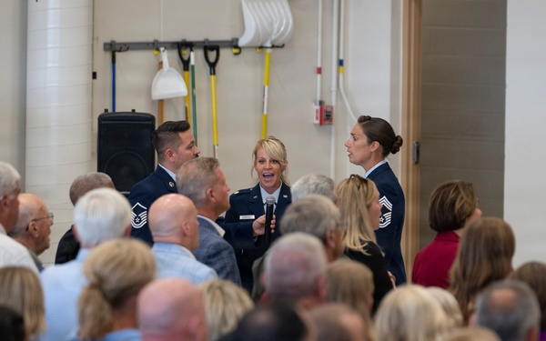 A military ceremony or event is taking place indoors, likely in a gymnasium or multipurpose facility. Three uniformed personnel, appearing to be U.S. Air Force members based on their dark blue dress uniforms, are addressing a large crowd of civilian attendees. The central figure, a female officer, is holding a microphone and appears to be speaking or presenting.