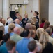 A military ceremony or event is taking place indoors, likely in a gymnasium or multipurpose facility. Three uniformed personnel, appearing to be U.S. Air Force members based on their dark blue dress uniforms, are addressing a large crowd of civilian attendees. The central figure, a female officer, is holding a microphone and appears to be speaking or presenting.