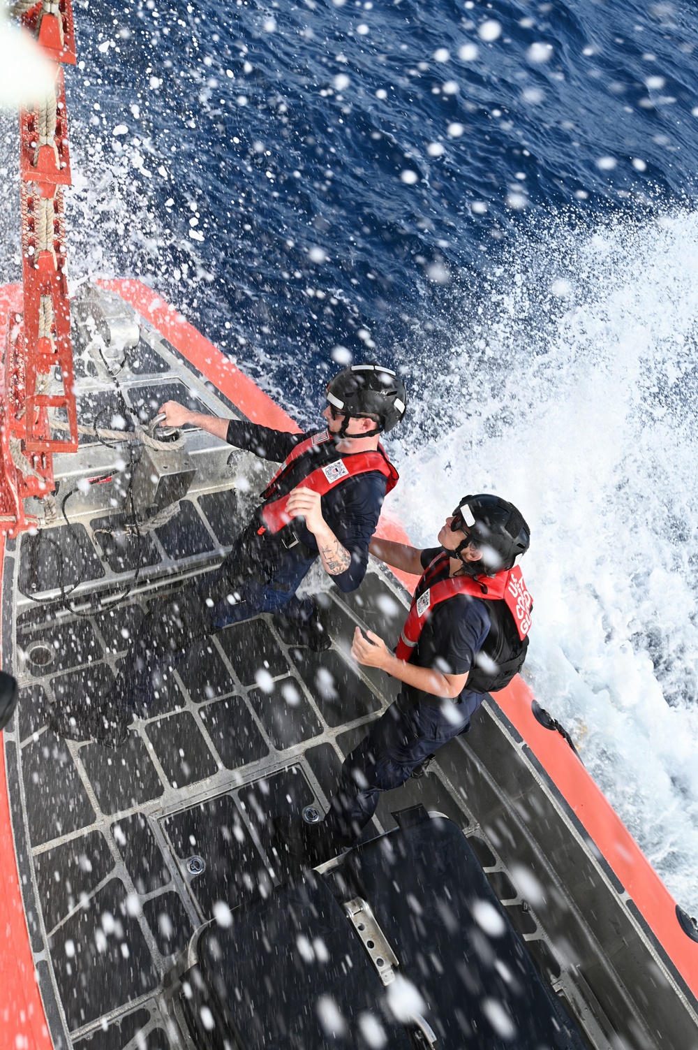 Two U.S. Coast Guard personnel are positioned on the deck of a vessel moving at speed through open ocean water. Both individuals are wearing red Coast Guard life vests, black helmets, and dark uniforms, and are being drenched by sea spray. The image is taken from an overhead angle, capturing the dramatic conditions of the operation.