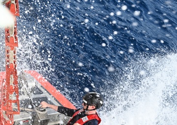Two U.S. Coast Guard personnel are positioned on the deck of a vessel moving at speed through open ocean water. Both individuals are wearing red Coast Guard life vests, black helmets, and dark uniforms, and are being drenched by sea spray. The image is taken from an overhead angle, capturing the dramatic conditions of the operation.