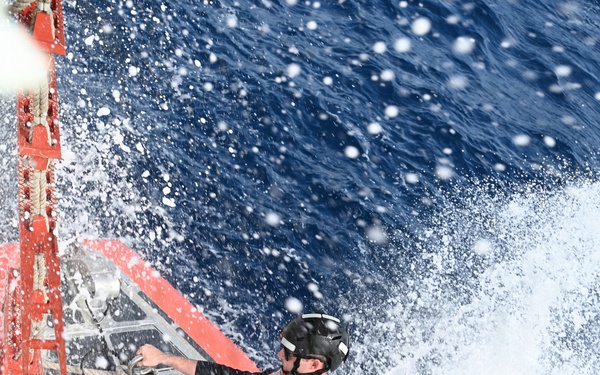 Two U.S. Coast Guard personnel are positioned on the deck of a vessel moving at speed through open ocean water. Both individuals are wearing red Coast Guard life vests, black helmets, and dark uniforms, and are being drenched by sea spray. The image is taken from an overhead angle, capturing the dramatic conditions of the operation.