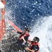Two U.S. Coast Guard personnel are positioned on the deck of a vessel moving at speed through open ocean water. Both individuals are wearing red Coast Guard life vests, black helmets, and dark uniforms, and are being drenched by sea spray. The image is taken from an overhead angle, capturing the dramatic conditions of the operation.