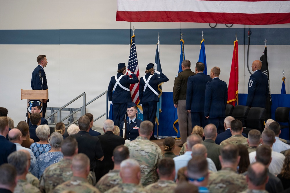 A formal military ceremony is taking place in what appears to be a hangar or gymnasium, likely a change of command or activation ceremony. A color guard is presenting flags while senior military personnel stand at attention on a stage, with a large audience of uniformed and civilian attendees seated in the foreground. An American flag is prominently displayed as a backdrop along with multiple military branch flags.