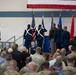 A formal military ceremony is taking place in what appears to be a hangar or gymnasium, likely a change of command or activation ceremony. A color guard is presenting flags while senior military personnel stand at attention on a stage, with a large audience of uniformed and civilian attendees seated in the foreground. An American flag is prominently displayed as a backdrop along with multiple military branch flags.