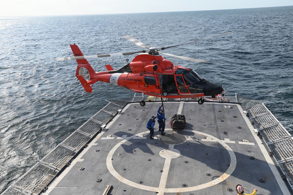 A U.S. Coast Guard helicopter, painted in distinctive international orange, hovers over the flight deck of a Coast Guard cutter while two crew members on deck handle a cargo load suspended by a hoist line. The operation appears to be a vertical replenishment or equipment transfer exercise conducted at sea. The helicopter is identifiable as an MH-65 Dolphin, a common Coast Guard rotary-wing aircraft.