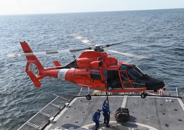 A U.S. Coast Guard helicopter, painted in distinctive international orange, hovers over the flight deck of a Coast Guard cutter while two crew members on deck handle a cargo load suspended by a hoist line. The operation appears to be a vertical replenishment or equipment transfer exercise conducted at sea. The helicopter is identifiable as an MH-65 Dolphin, a common Coast Guard rotary-wing aircraft.