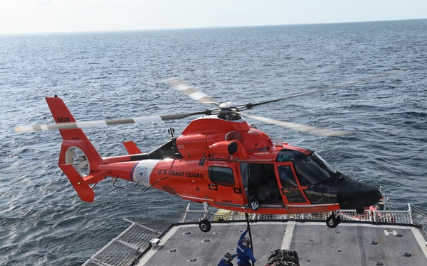 A U.S. Coast Guard helicopter, painted in distinctive international orange, hovers over the flight deck of a Coast Guard cutter while two crew members on deck handle a cargo load suspended by a hoist line. The operation appears to be a vertical replenishment or equipment transfer exercise conducted at sea. The helicopter is identifiable as an MH-65 Dolphin, a common Coast Guard rotary-wing aircraft.
