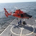 A U.S. Coast Guard helicopter, painted in distinctive international orange, hovers over the flight deck of a Coast Guard cutter while two crew members on deck handle a cargo load suspended by a hoist line. The operation appears to be a vertical replenishment or equipment transfer exercise conducted at sea. The helicopter is identifiable as an MH-65 Dolphin, a common Coast Guard rotary-wing aircraft.