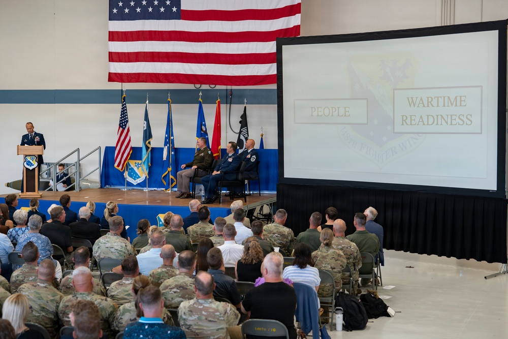 A military ceremony or briefing is taking place in a large hangar or assembly hall, with a speaker at a podium addressing a seated audience of military personnel and civilians. A large projection screen displays the words 'PEOPLE' and 'WARTIME READINESS,' suggesting a command briefing or change of command ceremony. Several officers are seated on stage beneath a large American flag and multiple military unit flags.