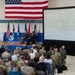 A military ceremony or briefing is taking place in a large hangar or assembly hall, with a speaker at a podium addressing a seated audience of military personnel and civilians. A large projection screen displays the words 'PEOPLE' and 'WARTIME READINESS,' suggesting a command briefing or change of command ceremony. Several officers are seated on stage beneath a large American flag and multiple military unit flags.