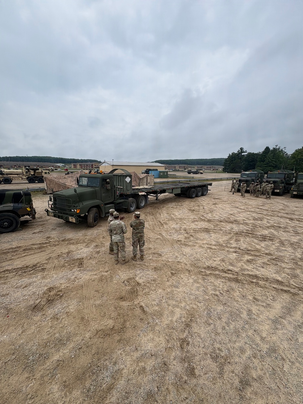A military training area showing soldiers in camouflage uniforms observing a large military flatbed truck and trailer on a dirt staging area. Multiple military vehicles are visible across the compound, with military installation buildings and tree lines visible in the background. The scene appears to depict a vehicle movement or logistics training exercise.