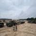A military training area showing soldiers in camouflage uniforms observing a large military flatbed truck and trailer on a dirt staging area. Multiple military vehicles are visible across the compound, with military installation buildings and tree lines visible in the background. The scene appears to depict a vehicle movement or logistics training exercise.