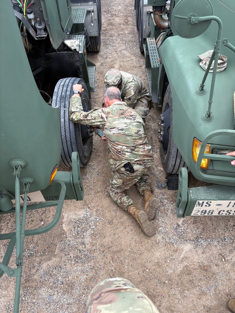 Two soldiers in OCP (Operational Camouflage Pattern) uniforms are performing maintenance or mechanical work on military vehicles, with one soldier lying prone on the ground and another crouching beside him. They appear to be working on the undercarriage or wheel assembly area between two olive-drab military trucks. A third soldier's helmet is partially visible at the bottom of the frame.