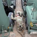 Two soldiers in OCP (Operational Camouflage Pattern) uniforms are performing maintenance or mechanical work on military vehicles, with one soldier lying prone on the ground and another crouching beside him. They appear to be working on the undercarriage or wheel assembly area between two olive-drab military trucks. A third soldier's helmet is partially visible at the bottom of the frame.